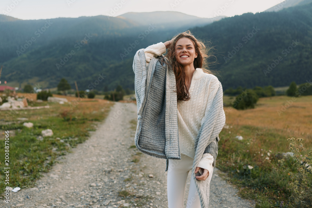 Fototapeta premium Smiling woman in cozy sweater and shawl walking on rural path with mountains in background. Outdoor lifestyle, nature, happiness, and casual fashion concept.