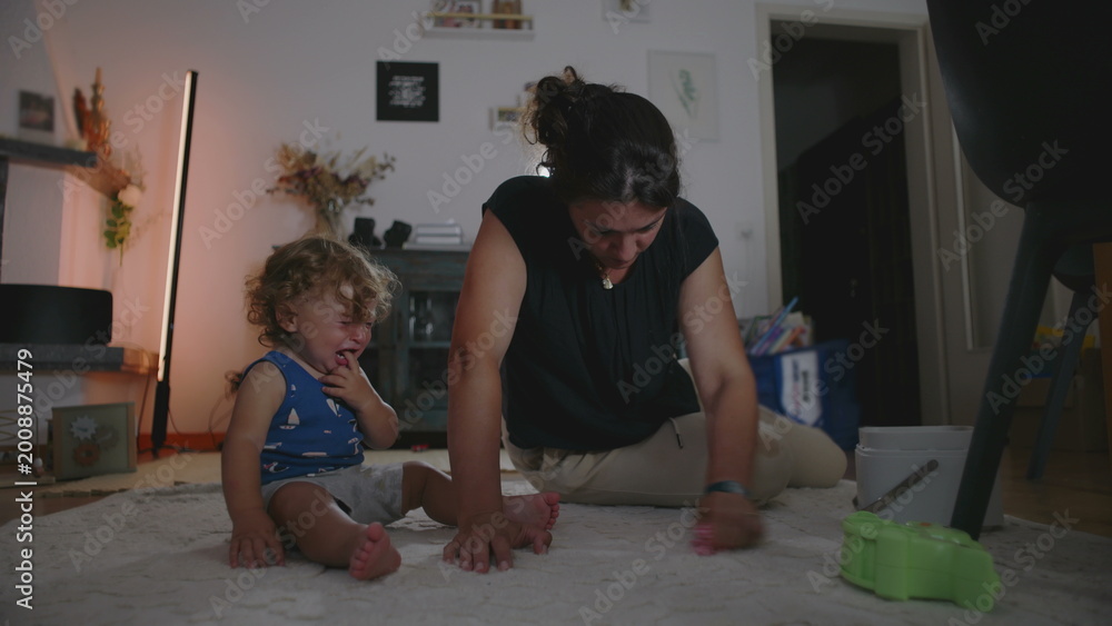 Naklejka premium Mother rubbing and cleaning carpet to remove a stain while toddler cries beside her seeking attention, candid indoor family moment showing daily parenting challenges
