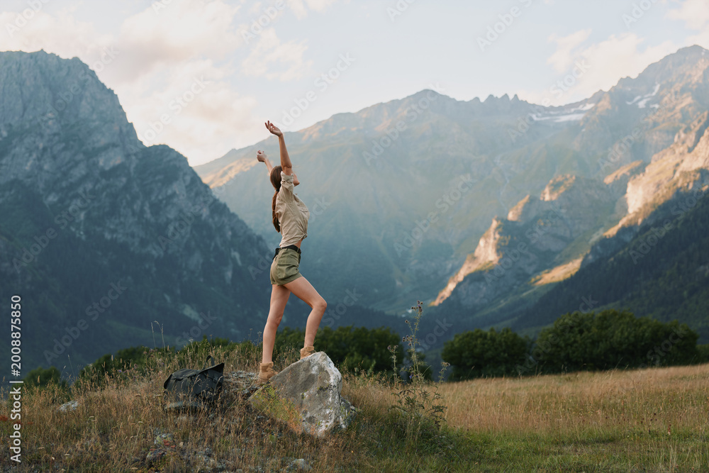 Fototapeta premium Young woman in casual clothes stands on a rock with arms raised in a mountain field, enjoying freedom and nature during a sunny day.