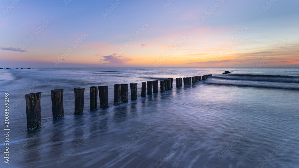 Fototapeta premium Wooden groynes in the sea at sunset – North Sea coast, Schoorl, the Netherlands. A line of weathered wooden groynes stretches from the foreground into the sea.