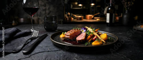 A fine dining main course of seared roasted sliced meat with seasonal vegetables, herb garnish and reduction sauce, paired with red wine on a dark moody restaurant table.