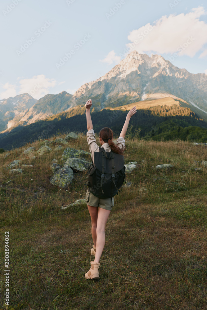 Fototapeta premium Smiling woman with a backpack raising her arms in a grassy meadow, mountains in the background. Outdoor lifestyle, adventure, and personal achievement. Bright natural light.