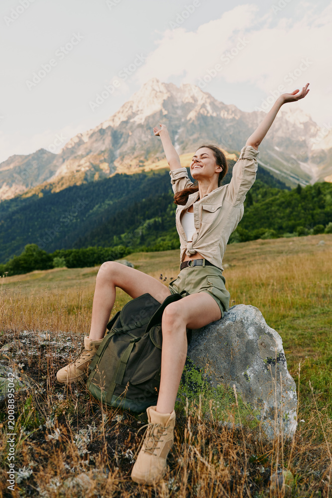 Fototapeta premium Happy woman in casual clothes sitting on a rock with arms raised in a mountain field. Concept of lifestyle, freedom, nature, and outdoor adventure. Bright natural light.