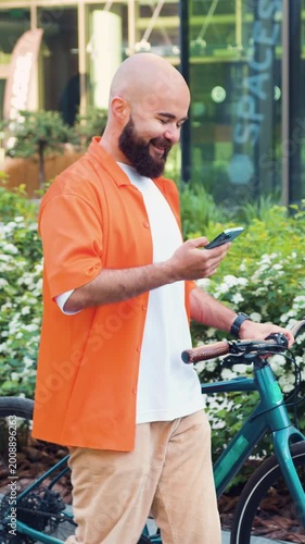 Caucasian man using smartphone typing a message while being leaned on the bicycle on modern city building background. Bicycle in city public eco transport mobile application walking on urban city.