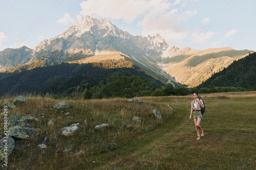 Naklejka premium Hiking in a meadow with mountains in the distance, wearing a backpack and trekking gear. A scenic landscape captures outdoor lifestyle, nature, and calm adventure.