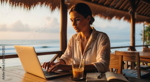 A young woman working on her laptop at a tropical beach cafe during a beautiful golden hour sunset, representing the digital nomad lifestyle and remote work freedom.