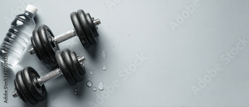 Pair of black dumbbells and a plastic water bottle with water droplets on a light grey background, fitness concept.