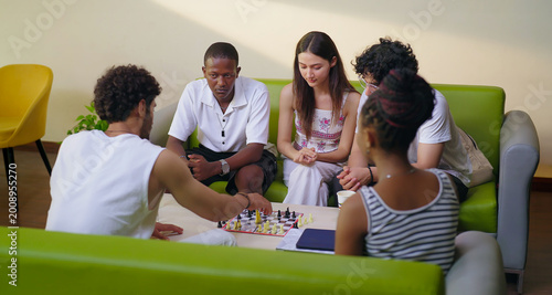 Indian young adult gen z male female group team sitting on sofa couch play chess indoor room place teen girl boy look game watch match talk speak share idea spend break time enjoy new day have fun joy