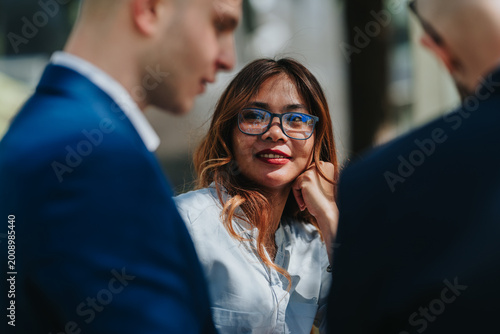 Fotografija A smiling businesswoman wearing glasses listens attentively while colleagues talk