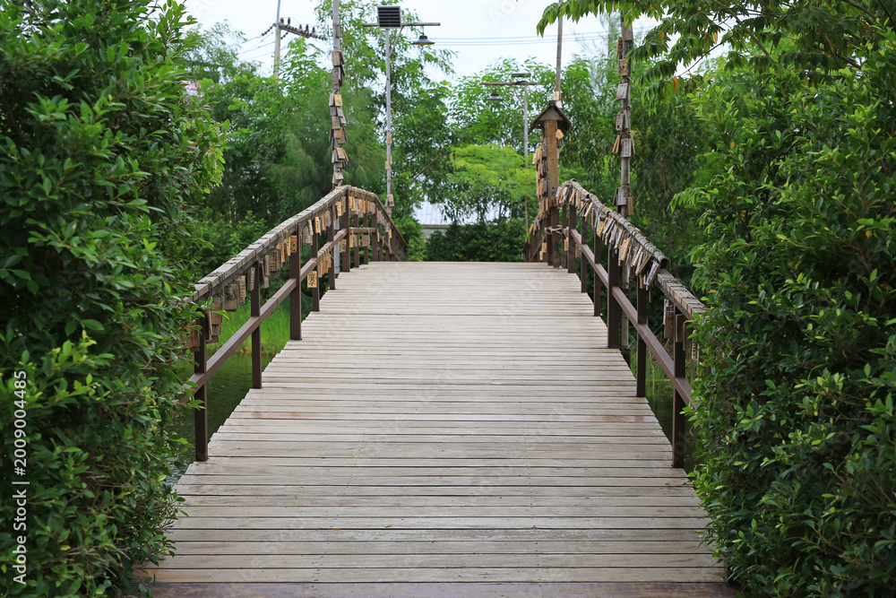 Fototapeta premium A peaceful wooden footbridge stretches across a quiet pond, flanked by lush green bushes and decorated with traditional hanging wooden tags.