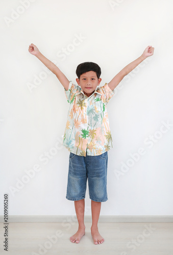 Cheerful young Asian boy in tropical palm tree print shirt and denim shorts standing against white wall with his arms raised high in a celebration pose.