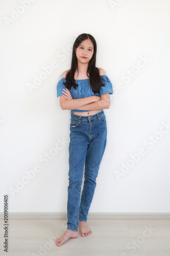 Portrait of young Asian girl in stylish denim outfit standing confidently and looking at camera against white wall background.