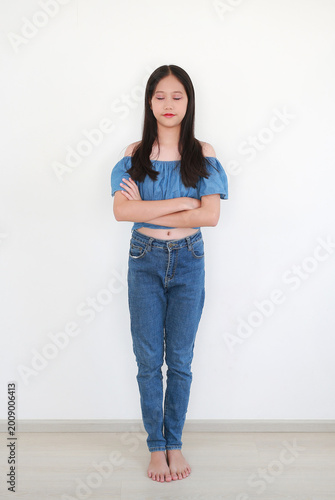 Young Asian girl in blue off-the-shoulder top and denim jeans standing with her arms crossed and eyes closed against white wall background.