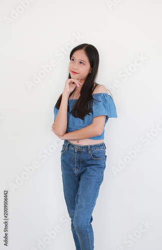 Young girl in blue off-the-shoulder denim top and matching jeans, stands on white background, resting her chin on her hand and looking upward with a curious expression.