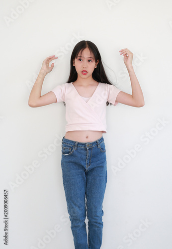 Portrait of young Asian girl in pink top and blue jeans posing playfully while looking beside against white background.