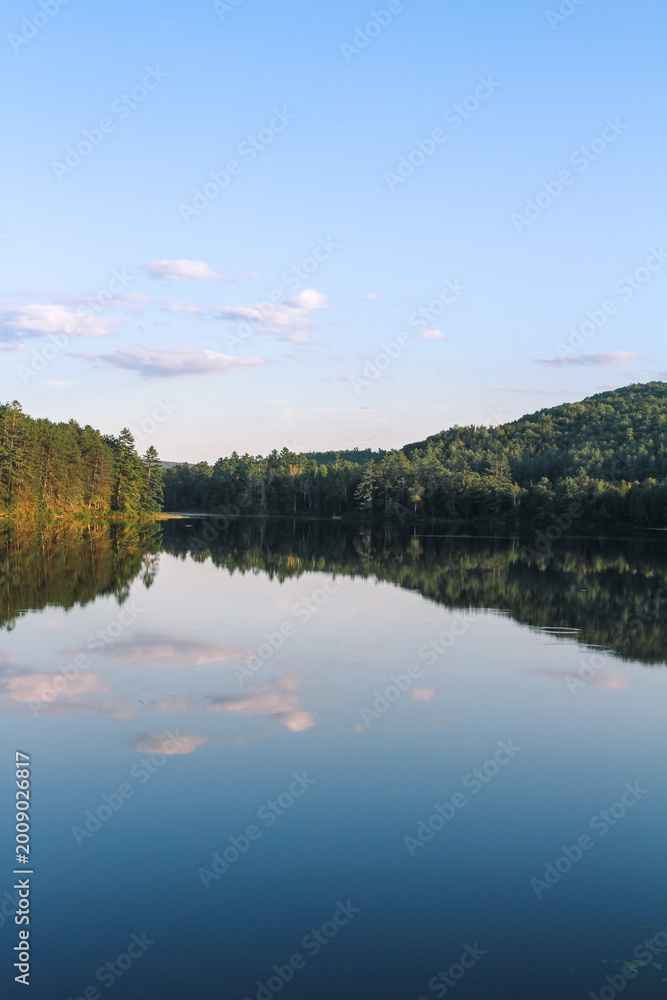 Obraz premium Glassy water reflects forested hills and soft clouds at Ogontz Lake in New Hampshire’s White Mountains near Camp Ogontz at golden hour.