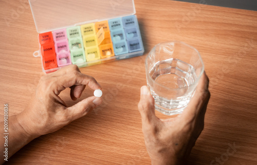 Senior hands holding pill and glass of water on wooden table with medicine box and blister packs, home healthcare and medication routine concept with copy space for text or image.