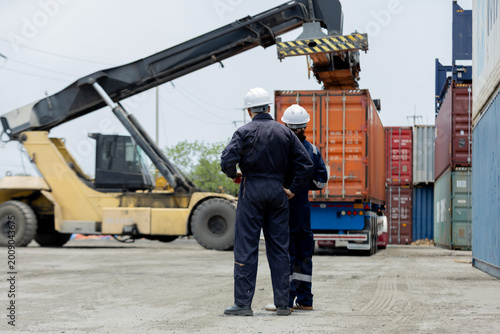 Bilde på lerret Two workers in protective gear oversee a container being loaded by heavy machinery at a shipping yard