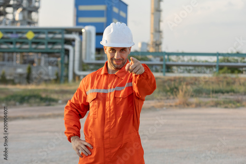 Fotografía A confident industrial worker in an orange coverall and white helmet points forward at a processing plant or refinery