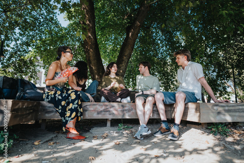 Bild auf Leinwand A group of young friends sit and talk on a wooden bench under shady trees