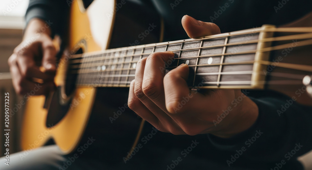 Fototapeta premium Close-up of a person playing an acoustic guitar, fingers positioned on the fretboard, natural light illuminating the scene indoors