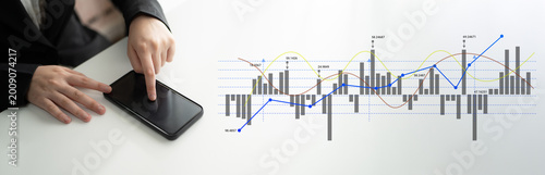 Fényképezés A businessman interacts with a smartphone displaying financial data while analyzing stock market trends on a nearby screen, showcasing modern business technology and analysis