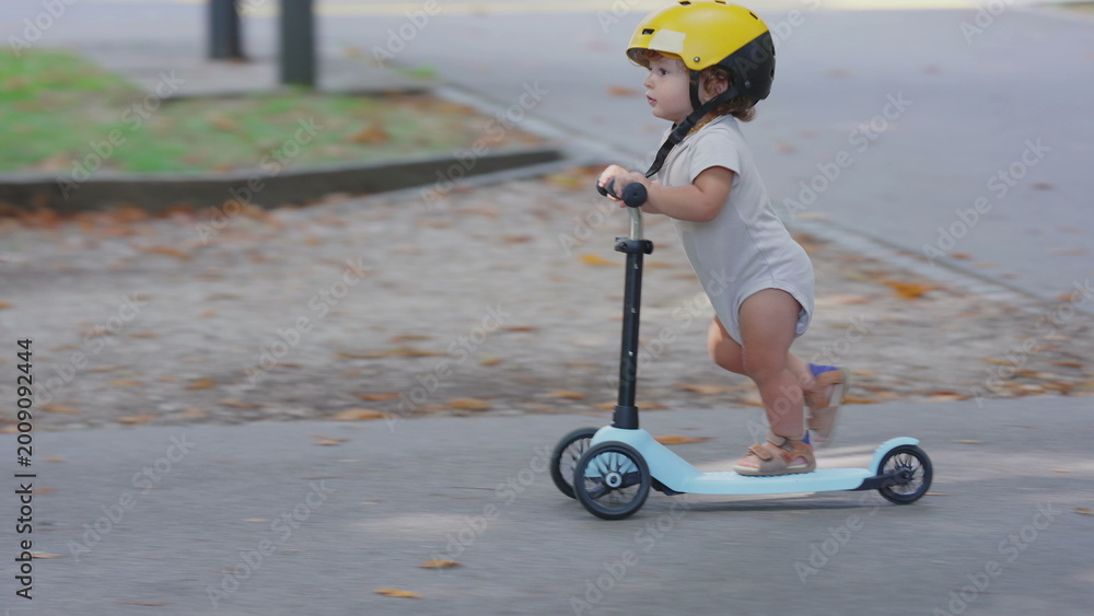 © Marco - Toddler wearing yellow helmet riding blue scooter in park pathway, captured in side profile, focused and balanced while gliding forward during playful outdoor moment