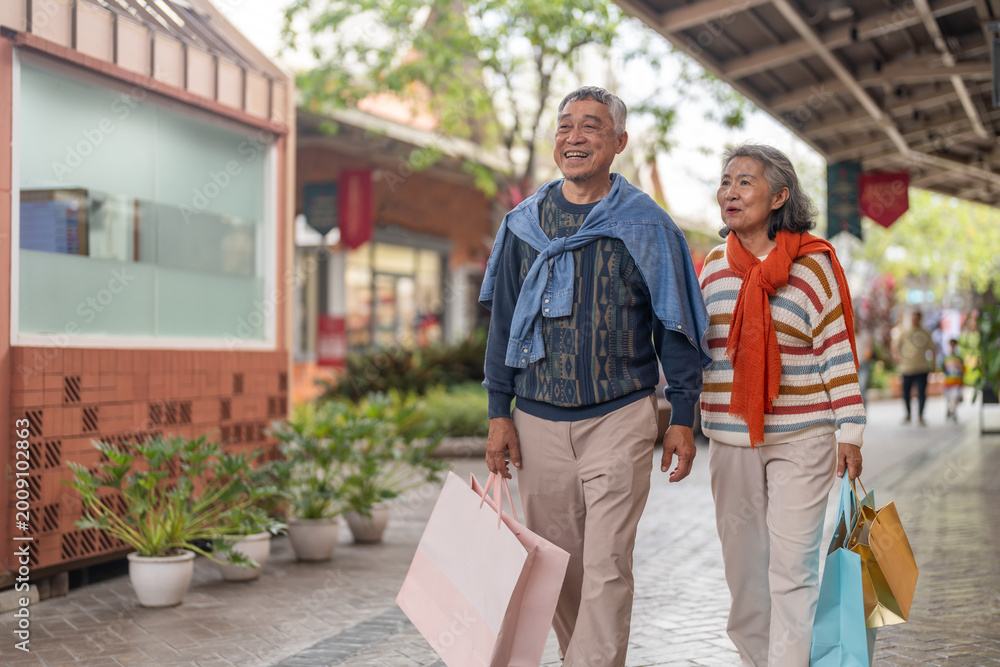 Naklejka premium Senior asian couple walking with shopping bags at mall smiling and laughing, love retirement lifestyle, shopping happiness and active senior living, purchase sale promotion, retail therapy