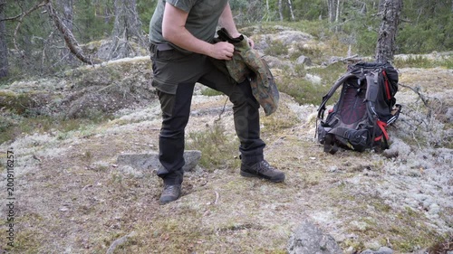 A hiker organizes outdoor clothing and gear near a large backpack on mossy rocky ground in a quiet boreal forest wilderness.