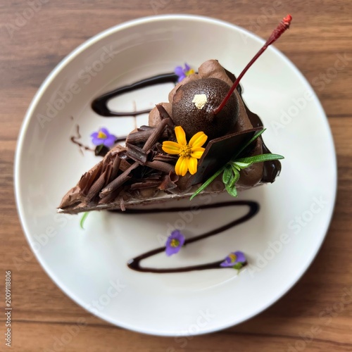 Quadro su tela Top view of a decadent chocolate mousse cake slice decorated with chocolate shavings, edible gold leaf, and vibrant flowers on a white plate over a wooden background