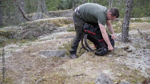 A male hiker adjusts his backpack on rocky moss-covered ground in a quiet conifer forest, preparing for outdoor trekking and wilderness travel.