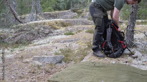 Outdoor adventurer prepares gear beside a backpack and groundsheet in a quiet pine forest campsite, surrounded by moss, rocks, and wilderness terrain.