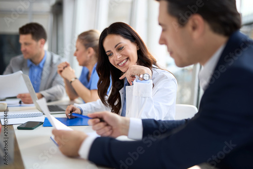 Young female doctor cooperating with an entrepreneur while going through paperwork during a meeting with their colleagues in the office
