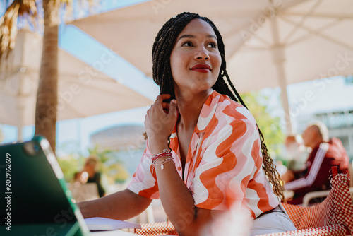 Fotografia Smiling woman with braided hair sitting at table beside tablet, symbolizing confidence, optimism, and the role of mobile technology in empowering modern freelance and digital lifestyle