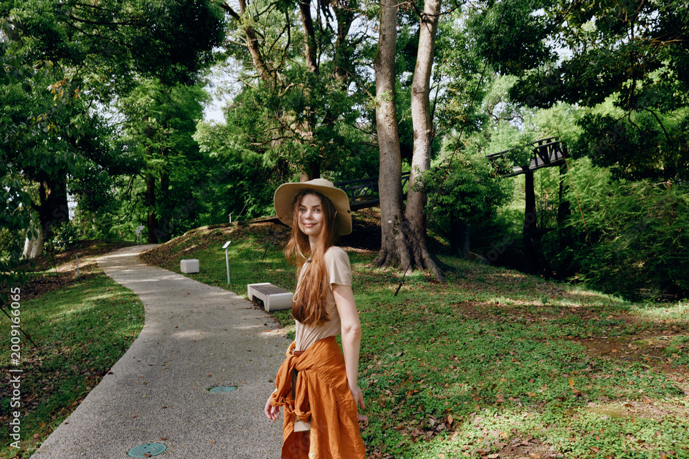 Fototapeta premium Woman walking in park on gravel path among tall trees, wearing a wide hat and orange skirt, smiling at camera. Outdoor nature portrait, summer leisure and relaxed lifestyle scene.