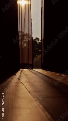 Vertical video. Sliding Curtains Opening to Large Floor to Ceiling Window with Warm Morning Sunlight Casting Long Shadows Across Wooden Floor