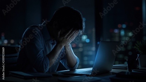 A man sits at a laptop at night with his head in his hands, expressing stress, burnout, overwork, and mental fatigue in modern work life.