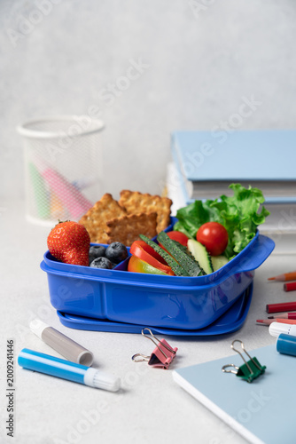 Blue school lunch box with berries crackers and vegetables on light background, side view with stationery, back to school concept