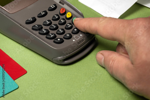 A hand is pressing buttons on a payment terminal placed on a green surface. Nearby are several credit cards. This scene takes place in a retail environment