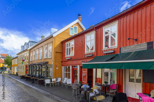 Bild auf Leinwand A street view of historic colorful wooden buildings in Trondheim, Norway, featuring red and yellow facades with outdoor cafe seating under a bright blue sky