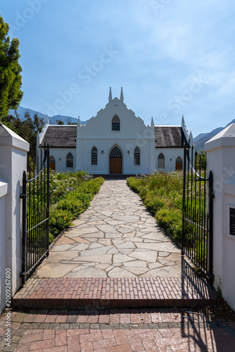 A charming church in Franschhoek under a bright blue sky, surrounded by mountains and vineyards, glowing in warm sunlight with a relaxed holiday atmosphere and visiting tourists.