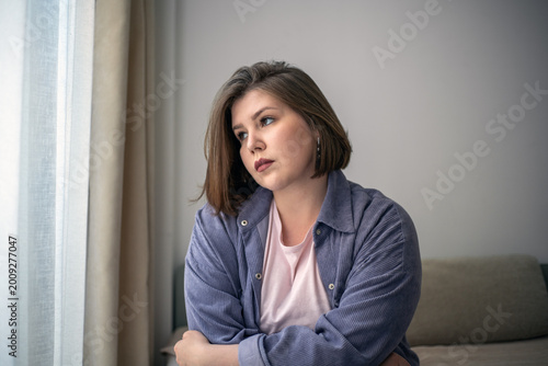 Thoughtful plus size woman looking through window, sad home portrait, self reflection, mental health, emotional wellness, inclusive lifestyle.