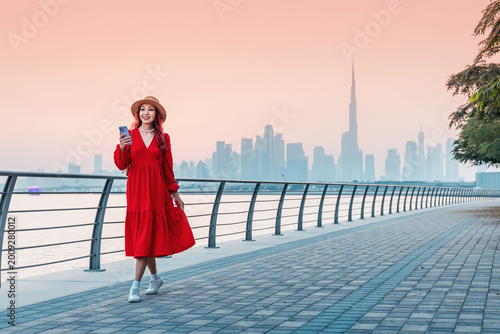 Young woman in red dress enjoying a walk along the waterfront promenade, holding a phone with the iconic Dubai skyline