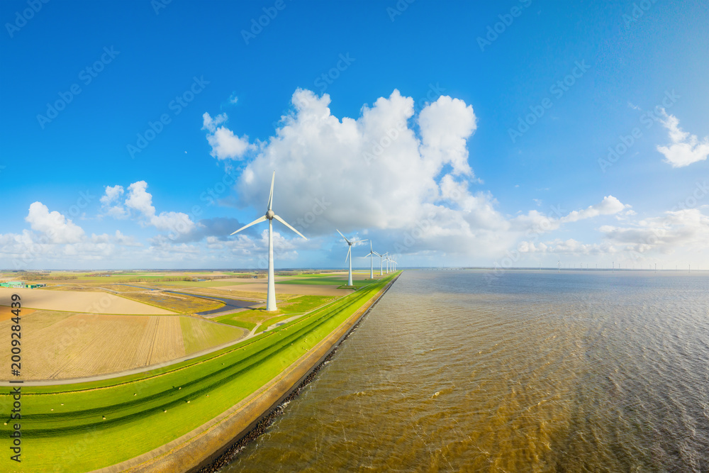 Fototapeta premium Aerial view of wind turbines on green fields near sea coast and dam. Scenic landscape with wind generators, field and sky with clouds. Renewable energy and environment from above.