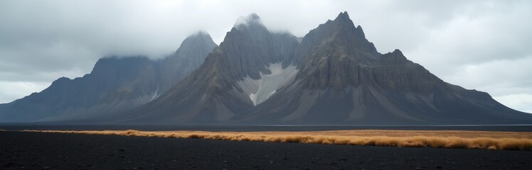 Jagged grey mountains with snow patches rise above dark volcanic sand. Golden grass borders the desolate landscape under a cloudy sky. This wild nature scene offers immense scale and solitude.