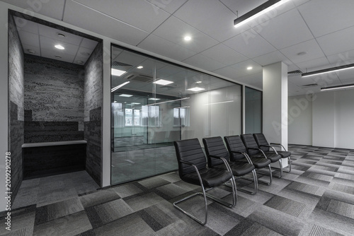 A corporate office features several chairs lined up in front of a glass meeting room. The design includes clean lines and a neutral color scheme. The area looks organized and ready for use