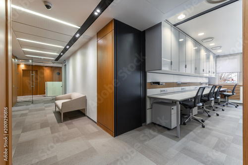 The scene presents a modern office layout with a seating area on one side and workstations on the other. Large windows allow natural light to fill the space