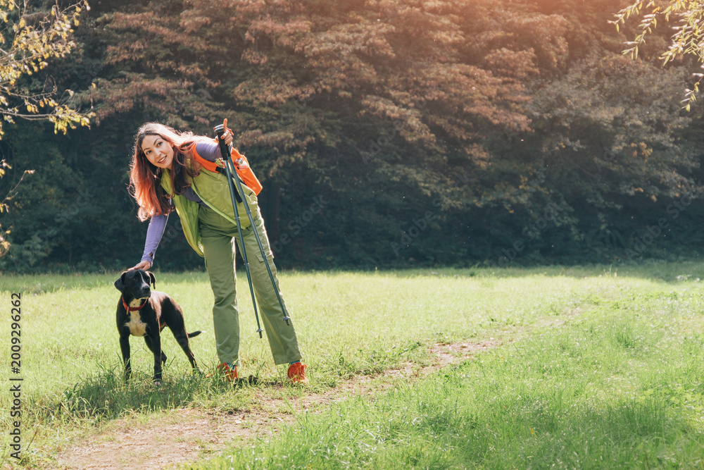 Fototapeta premium Happy woman wearing backpack and using trekking poles, petting her dog on a green grass outdoor trail, enjoying companionship