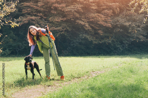 Happy woman wearing backpack and using trekking poles, petting her dog on a green grass outdoor trail, enjoying companionship