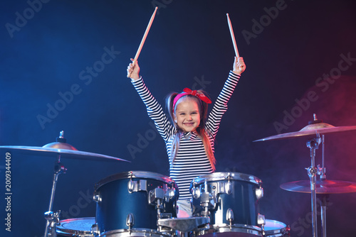Foto Smiling little girl in bandana learning how to play drums in color lights and sm